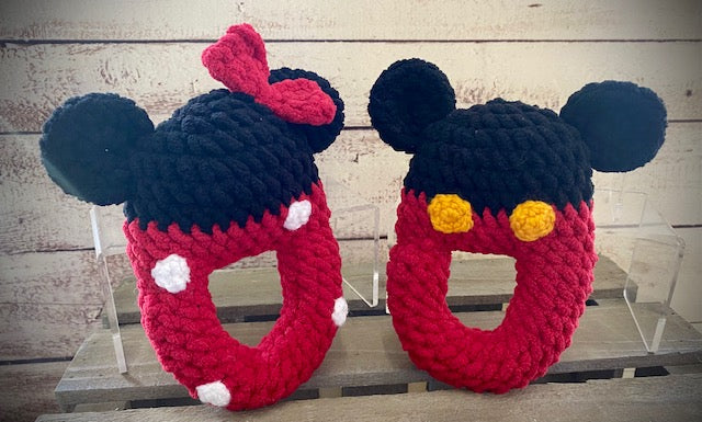 Two hand-crocheted rattle toys designed to resemble cartoon characters, one in colors red and black with a bow, and the other in colors black and red with a face detail.